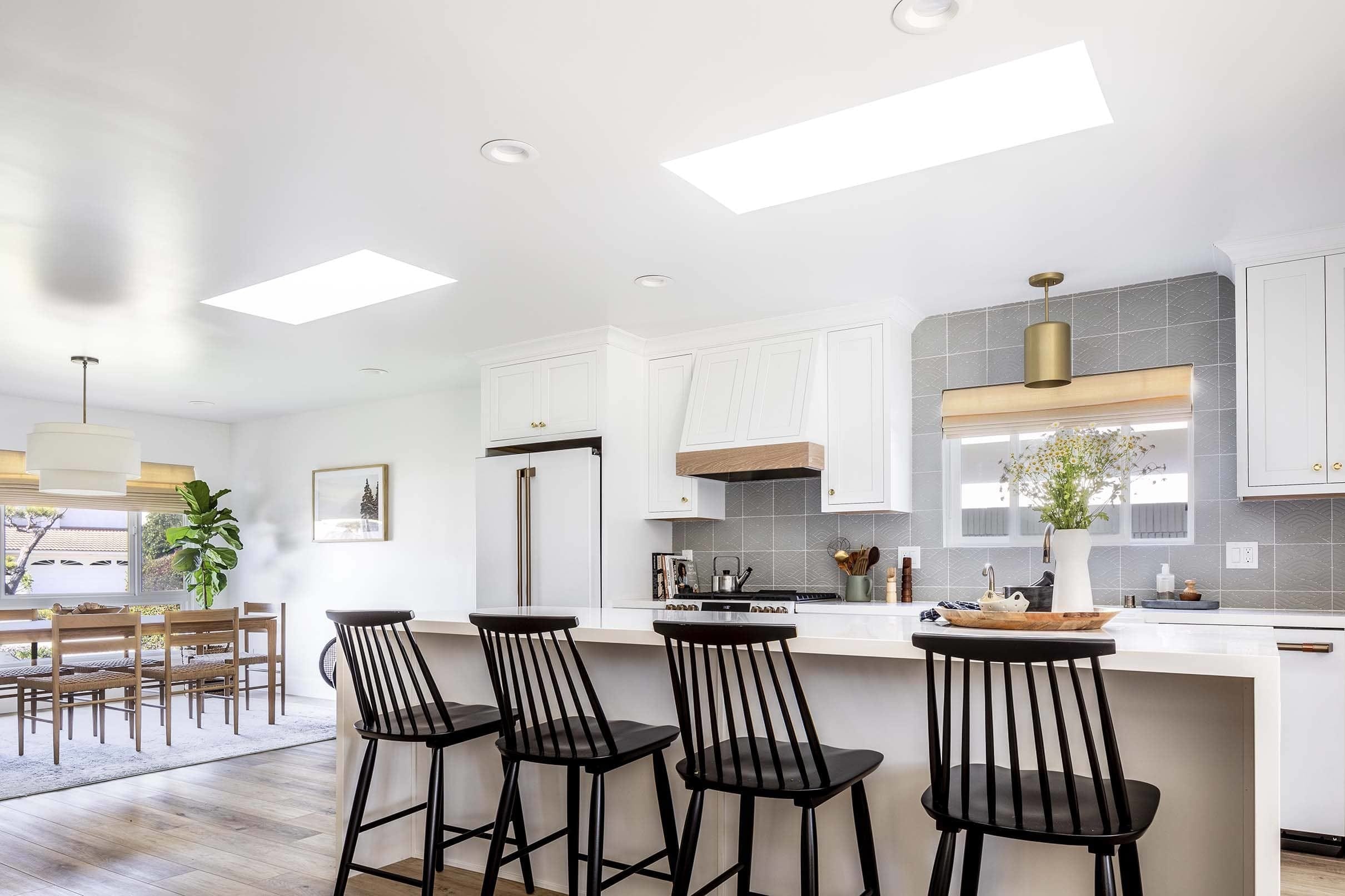 Two skylights brighten an all-white kitchen that includes white appliances, a grey backsplash, white waterfall island and black Shaker-style counter chairs