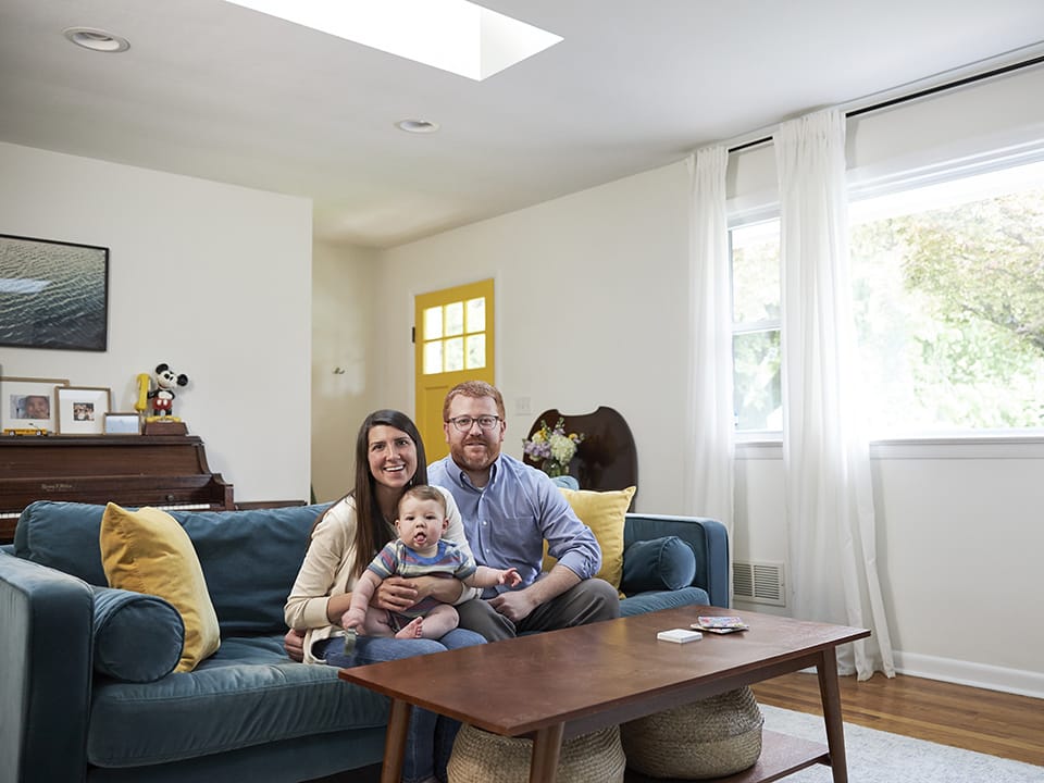 mom dad and infant sitting on a blue couch under a skylight in a living room