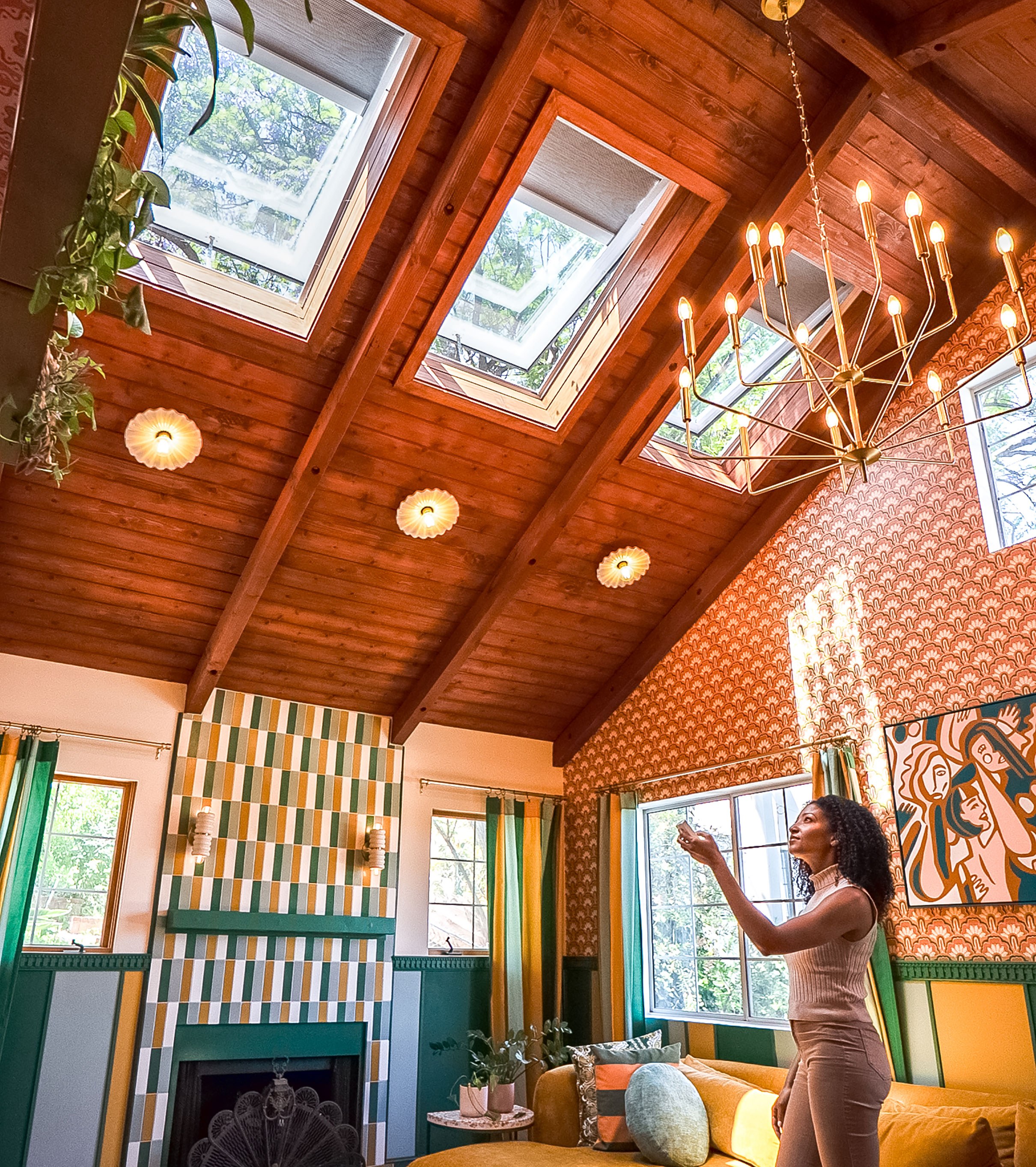 Photo taken at The Dazey Desert House, owned by influencer Dani Dazey, decorated in a maximalist style. There are three venting skylights in a wood-paneled roof in the open position with skylight shades halfway down. A model points a VELUX Touch Remote at the open Skylight Systems to begin closing the skylights and skylight shades.
