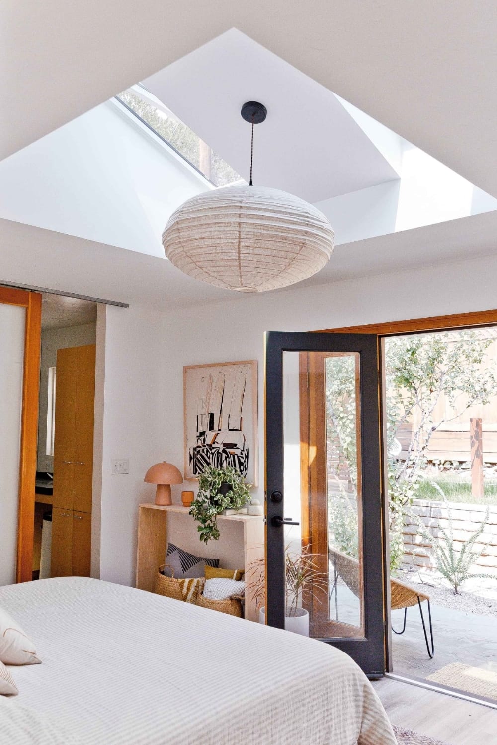A bedroom with two skylights with shades and a Japanese lantern light fixture mounted in between. Neutral color bedding and natural wood French door frame.