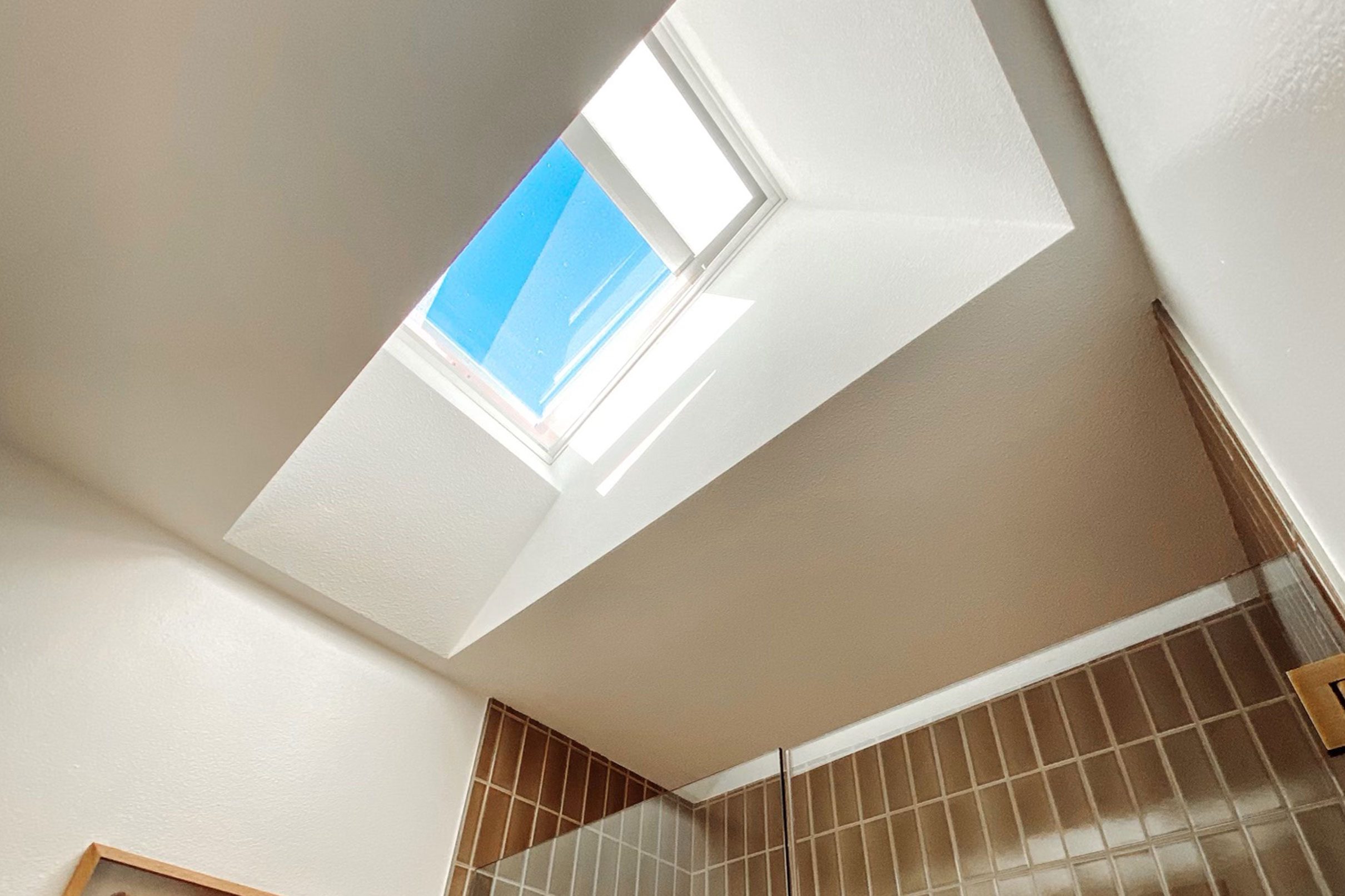 Angled view of a closed curb mount skylight with shade partially closed in a small bathroom with part of the beige tile wall shower visible.