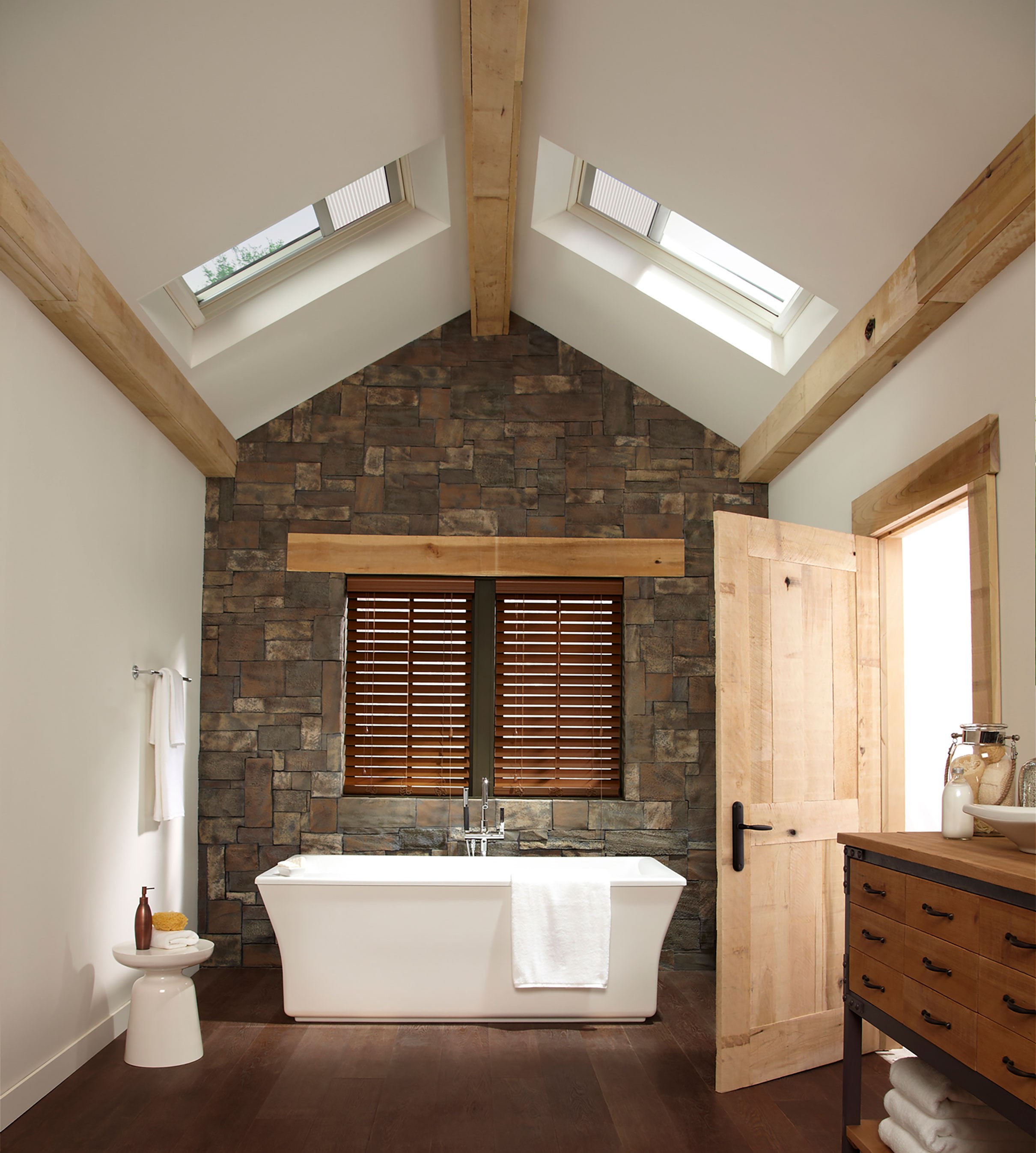 A modern bathroom with white standalone bathtub, stone accent wall, dark wooden floors, and light wooden accents, naturally lit by two fixed skylights with shades.