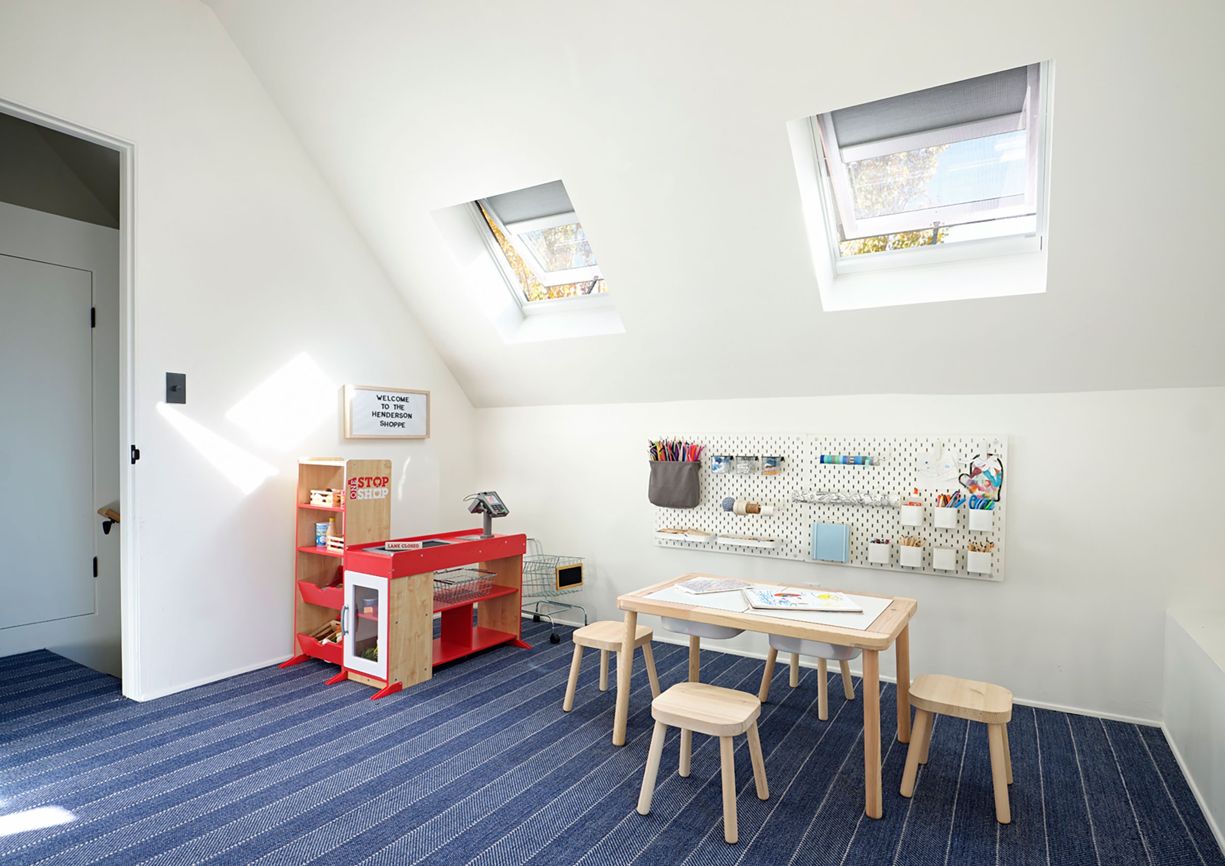 A kids play area with open skylights and gray shades partially lowered. The play area has a small desk with stools, a toy checkout counter, and crafting utensils hanging on the wall below the skylights.