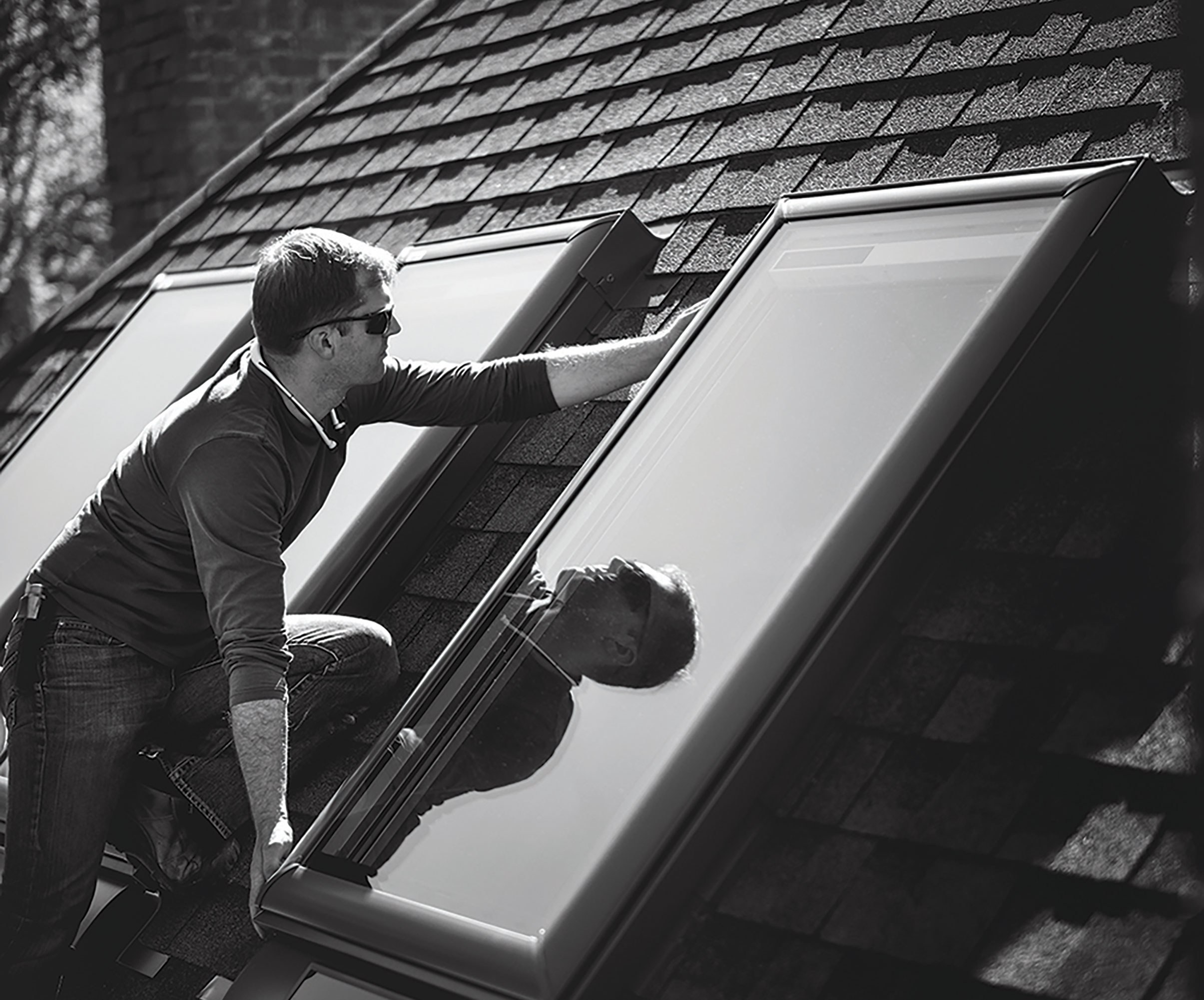 Black and white image of a skylight installer placing a curb-mounted skylight on a shingled roof exterior.