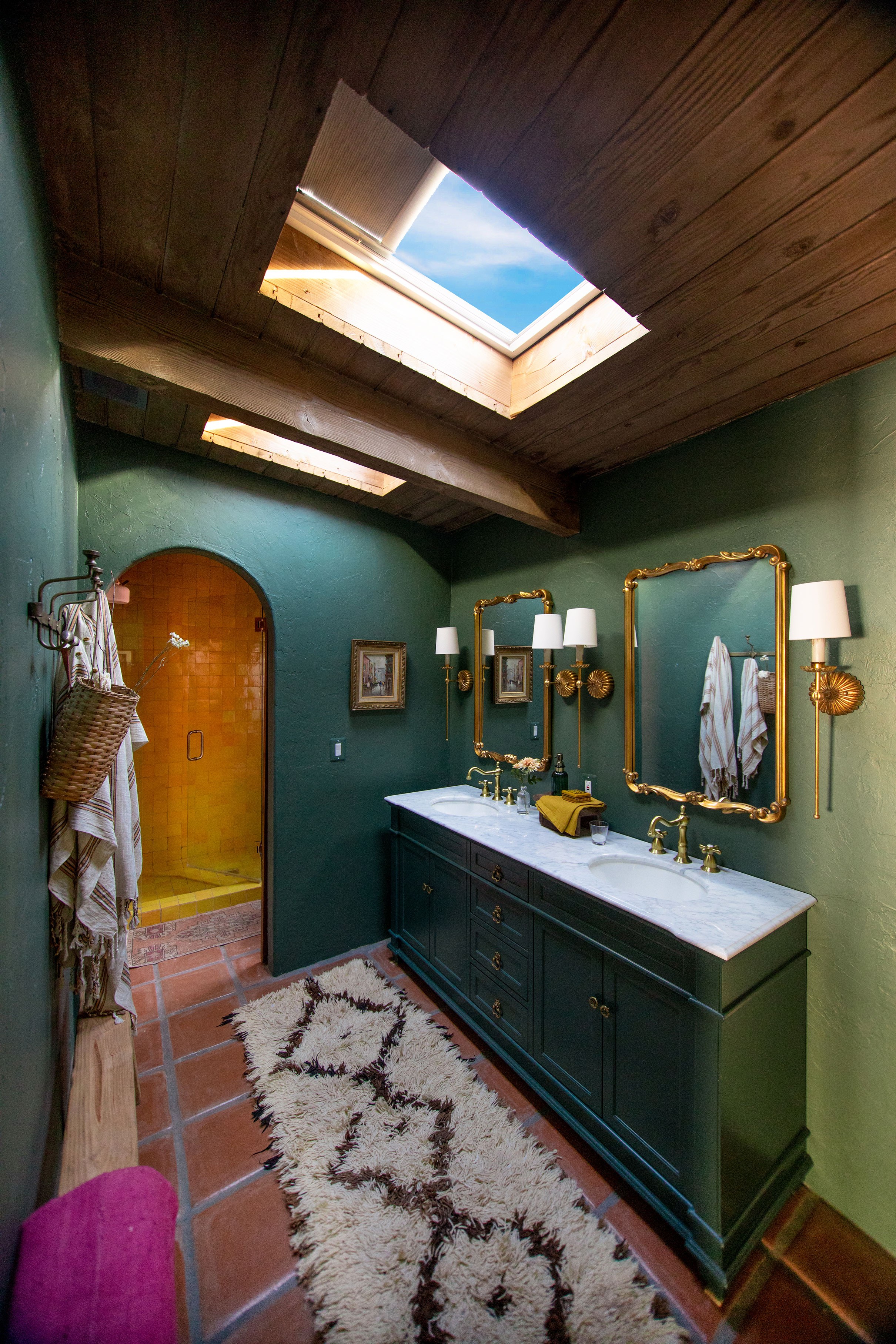 A bathroom renovation from Studio DIY! and Jeff Mindell in their Palm Springs home. The bathroom has two closed skylight systems with shades partially closed, deep green walls, brick flooring, and gold accents.